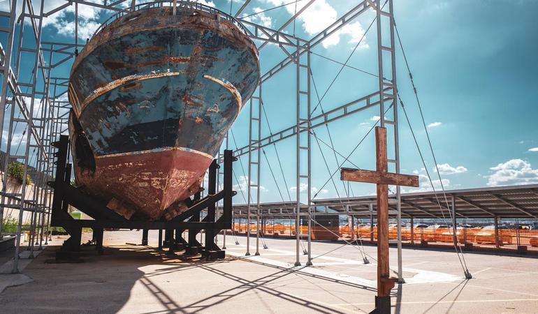 Augusta, 2024. Una croce in legno davanti al relitto del barcone affondato nel 2015 nel Mediterraneo, abbandonato sulla nuova darsena della città siciliana. Foto: Paolo Valenti