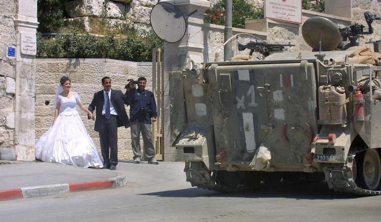 Betlemme (Cisgiordania), 7 luglio 2002. I novelli sposi Nader e Jalaa, abitanti del vicino campo profughi palestinese di Daheisheh, lasciano i giardini dell'ospedale della Sacra Famiglia, dove hanno fatto le foto del matrimonio, mentre un corazzato israeliano passa. Foto di Musa Al Shaer/Ansa