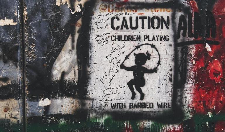 Una bambina salta la corda fatta di filo spinato, murales dell'artista Cakes Stencil su un muro in Palestina (Foto Jakob Rubner - Unsplash)